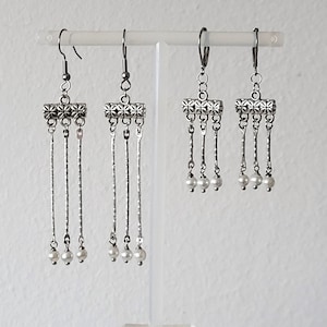 May include: Three pairs of silver dangle earrings with a decorative metal charm and three white pearl accents on each earring.