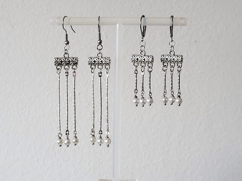 May include: Three pairs of silver dangle earrings with a decorative metal charm and three white pearl accents on each earring.