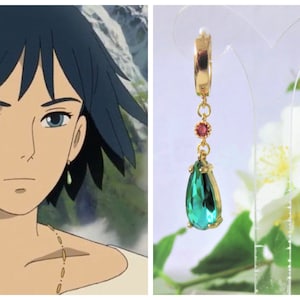 May include: A pair of gold hoop earrings with a dangling teardrop-shaped green gemstone and a small red gemstone accent. The earrings are on a white background with white flowers.