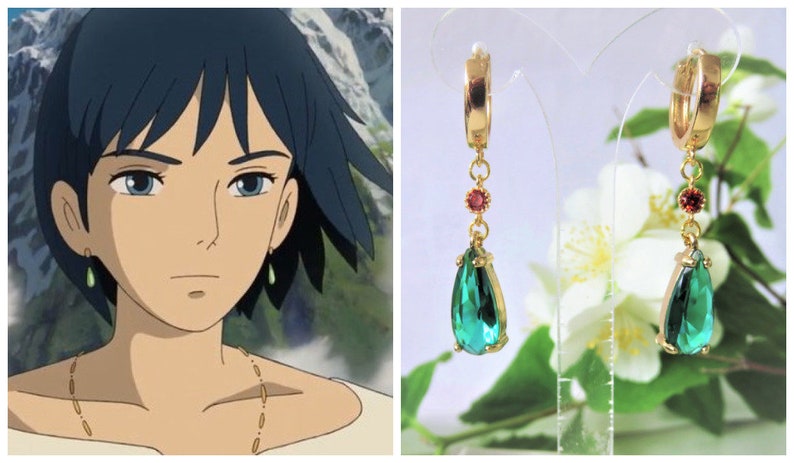 May include: A pair of gold hoop earrings with a dangling teardrop-shaped green gemstone and a small red gemstone accent. The earrings are on a white background with white flowers.