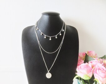 May include: A silver layered necklace with a round pendant and small dangling charms. The necklace is displayed on a black mannequin bust.