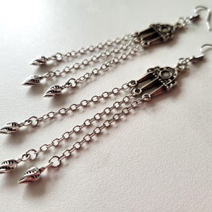 Peut inclure: Boucles d'oreilles pendantes argentées avec des détails de filigrane complexes. Chaque boucle d'oreille présente un dessus décoratif avec trois chaînes, chacune se terminant par un charm en forme de feuille. Fermeture à crochet, sur fond blanc.