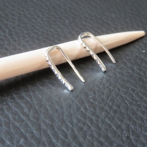 Könnte beinhalten: Ein Paar silberne Ohrringe mit einem strukturierten, rautenförmigen Design. Die Ohrringe sind gebogen und oben offen.