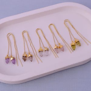 Puede incluir: Pendientes colgantes dorados con colgantes de piedras preciosas en varios colores, incluyendo morado, marrón, transparente y verde. Los pendientes se muestran en una bandeja ovalada blanca sobre un fondo azul claro. Las piedras preciosas tienen forma de cono.