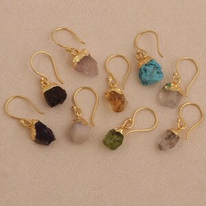 May include: A collection of gold-colored dangle earrings, each featuring a unique, irregularly shaped gemstone in various colors, including purple, green, and blue. The earrings are displayed on a neutral, light-colored surface.