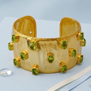 以下が含まれることがあります： 質感のあるブラッシュドメタル仕上げの幅広ゴールドトーンカフブレスレット。ブレスレットの外側には、個別のゴールドトーンのベゼルにセットされた、複数の楕円形でファセットカットされたペリドットカラーの緑色の石が装飾されています。