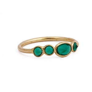 Puede incluir: Un anillo de oro con cinco piedras preciosas de color verde esmeralda. El anillo tiene una banda fina y continua, y las piedras preciosas son ovaladas y redondas, dispuestas en una fila. El anillo se muestra sobre un fondo blanco liso.
