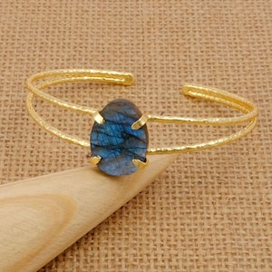 Op de afbeelding: Een goudkleurige manchetarmband met een getextureerde afwerking. De armband heeft een grote, ovaalvormige, blauwe labradorietsteen gezet in een gouden zetting. De steen heeft een iriserende kwaliteit. Sieraden.