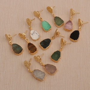 May include: A collection of gold-colored dangle earrings with various gemstone pendants. The earrings feature a textured, circular top and a teardrop-shaped gemstone in shades of green, black, white, brown, and pink. The earrings are displayed on a light beige surface.