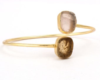 May include: A gold bangle bracelet with two square-shaped gemstones. The top gemstone is a light pink color, and the bottom gemstone is a light brown color.