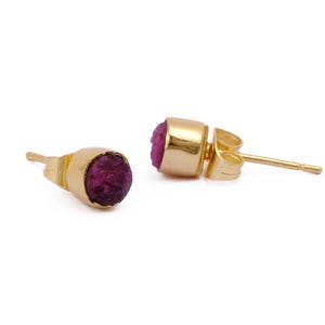 May include: A pair of gold stud earrings, each with a raw, deep pink gemstone set in a gold bezel. The earrings are presented against a white backdrop, accentuating the jewellery's design and colour. The earrings are suitable for pierced ears.