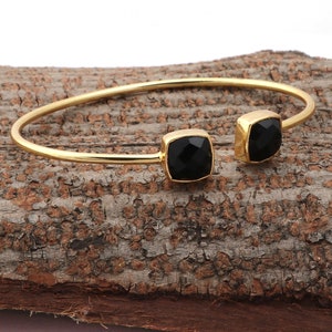 May include: A gold-coloured cuff bracelet featuring two square, black onyx stones. The open-ended bracelet is displayed on a textured, brown surface. The text "Black Onyx" is visible in the image.