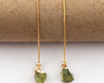 Puede incluir: Un par de pendientes colgantes dorados. Cada pendiente presenta una fina cadena dorada que sostiene una piedra preciosa verde oliva en bruto. Los pendientes se muestran sobre un fondo neutro.