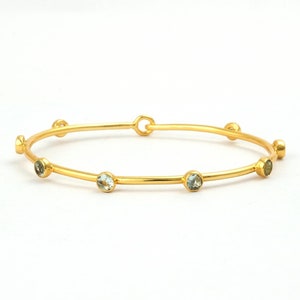 May include: A gold-colored bangle bracelet with six light blue gemstones evenly spaced around the band. The bracelet has a simple, elegant design and a clasp closure. The gemstones are round and add a touch of color to the piece.