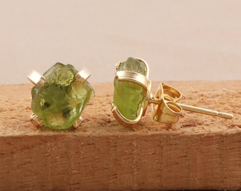 Puede incluir: Un par de pendientes de oro con piedras de peridoto verde claro en bruto. Las piedras de peridoto están sujetas por garras de oro y una banda de oro. Los pendientes se exhiben sobre una superficie de madera.