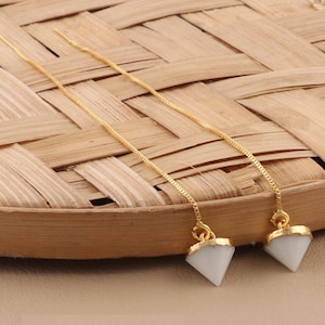 Può includere: Un paio di orecchini pendenti color oro con pendenti bianchi a forma di diamante. Gli orecchini sono esposti su una superficie intrecciata marrone chiaro.