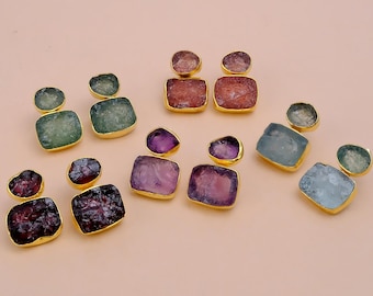 Puede incluir: Una colección de pendientes de piedras preciosas en varios colores, incluyendo verde, morado, rojo y azul. Cada pendiente tiene un engaste de color dorado. Las piedras preciosas tienen forma ovalada y cuadrada.