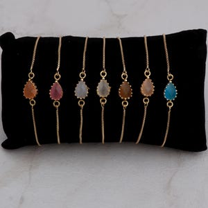 Puede incluir: Siete pulseras doradas con piedras preciosas en forma de lágrima en varios colores, como naranja, rosa, gris, amarillo, marrón y azul. Las pulseras se exhiben sobre una superficie de terciopelo negro.