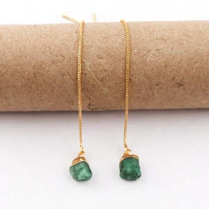 Puede incluir: Un par de pendientes colgantes dorados con piedras preciosas de color verde esmeralda. Los pendientes tienen un diseño de cadena delicada y se muestran sobre un fondo neutro.