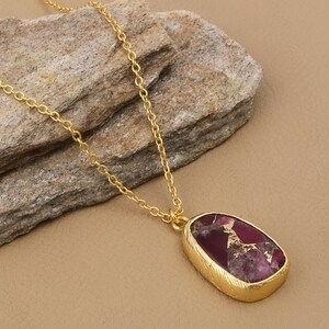 May include: A gold-colored necklace with a pendant featuring a maroon and gold stone. The pendant has a textured gold border and is suspended from a delicate gold chain. The necklace is displayed on a neutral-toned surface.