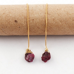 Puede incluir: Pendientes colgantes dorados con piedras preciosas de rubí rosa oscuro en bruto. Los pendientes presentan un delicado diseño de cadena, con las piedras de rubí en la parte inferior. El fondo es de color beige neutro.