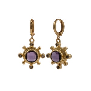 Puede incluir: Un par de pendientes de aro dorados. Cada pendiente presenta una piedra preciosa central, redonda y púrpura, rodeada de piedras preciosas claras más pequeñas y un diseño de sol. Los pendientes están diseñados con un cierre de bisagra.