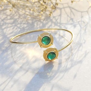 Puede incluir: Una pulsera abierta de tono dorado con dos engastes circulares, cada uno con una piedra verde esmeralda facetada. Los engastes están enmarcados por un borde metálico texturizado y punteado, creando un diseño equilibrado.