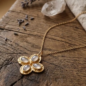 May include: A gold-colored necklace with a flower-shaped pendant featuring four oval, light-colored stones. The necklace rests on a rustic wooden surface, with dried lavender and a crystal nearby. The chain is delicate and the pendant has a textured border.