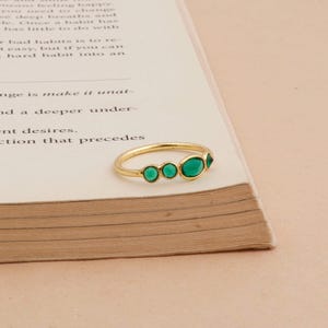 Puede incluir: Un anillo de oro con cinco piedras preciosas verdes. El anillo est&aacute; colocado sobre un libro con texto. Las piedras preciosas son ovaladas y triangulares, dispuestas en fila. Es una joya delicada.