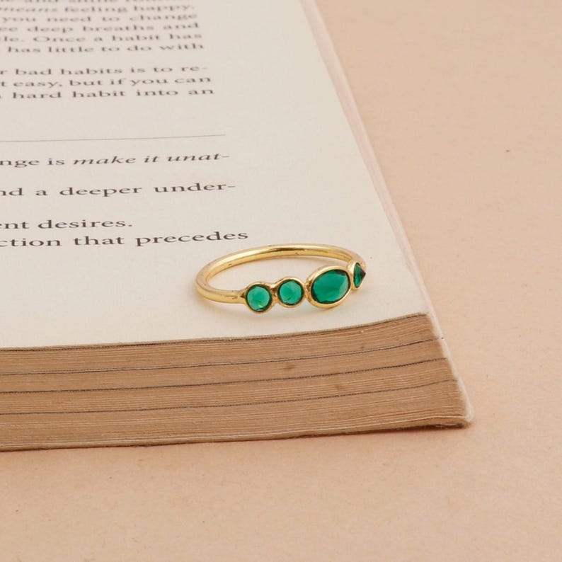 Puede incluir: Un anillo de oro con cinco piedras preciosas verdes. El anillo est&aacute; colocado sobre un libro con texto. Las piedras preciosas son ovaladas y triangulares, dispuestas en fila. Es una joya delicada.
