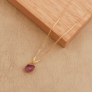 Op de afbeelding: Een goudkleurige ketting met een kleine, ruwe, roze edelsteen hanger. De hanger is onregelmatig van vorm en heeft een goudkleurige dop. De fijne ketting ligt op een lichtbruin oppervlak met een houten blok op de achtergrond.