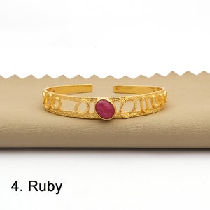 May include: A gold-colored cuff bracelet with an oval, ruby-colored stone in the center. The bracelet has a textured design with open circles along the sides. The text "4. Ruby" is at the bottom.