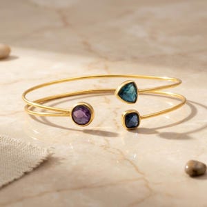 May include: Gold-tone open cuff bracelet with three gemstones. The bracelet features a round purple gemstone, a triangular teal gemstone, and a square dark blue gemstone. The bracelet is displayed on a light-colored surface.