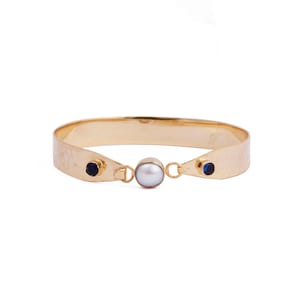 Peut inclure: Un bracelet doré avec une texture martelée. Il comporte une perle centrale et deux pierres précieuses bleu foncé. Le design est simple et élégant, adapté au quotidien ou aux occasions spéciales.