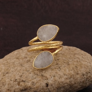 Op de afbeelding: Een goudkleurige ring met een spiraalvormig ontwerp. De ring heeft twee druppelvormige, lichtgrijze stenen. De ring wordt gepresenteerd op een lichtbruin, getextureerd oppervlak, met een donkerbruine achtergrond.