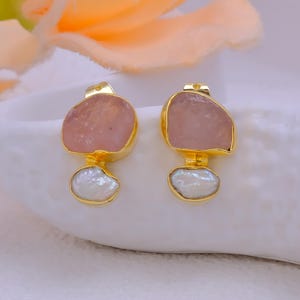 May include: A pair of gold-toned earrings featuring a large, irregular pink stone at the top and a white, pearlescent, teardrop-shaped stone below. The earrings are set against a soft, white background.
