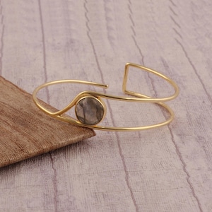 Puede incluir: Un brazalete abierto de color dorado con una piedra gris redonda en el centro. El brazalete tiene un diseño minimalista con una forma curva y un acento geométrico en un extremo. El brazalete se exhibe sobre una superficie de madera.