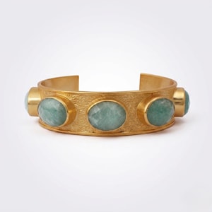 Könnte beinhalten: Goldfarbenes Armreif-Armband mit fünf ovalen, hellgrünen Edelsteinen. Das Armband hat eine strukturierte Oberfläche und ein offenes Design. Die Edelsteine sind gleichmäßig auf der Vorderseite angeordnet.