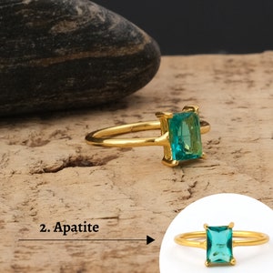 Puede incluir: Un anillo de oro con una piedra cuadrada de apatita verde esmeralda. El anillo está sobre una superficie de madera.