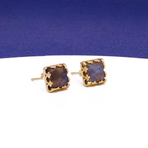 May include: A pair of square stud earrings with a gold-coloured filigree setting. Each earring features a square, iridescent, blue-grey stone. The earrings are displayed on a white surface with a blue background.
