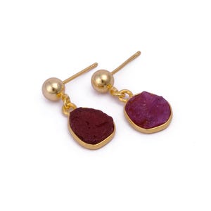 Peut inclure: Une paire de boucles d'oreilles à clous dorées avec une pierre précieuse rouge foncé de forme irrégulière. Les pierres précieuses sont serties dans une lunette dorée. Les boucles d'oreilles sont présentées sur fond blanc.