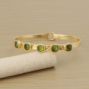 Op de afbeelding: Een goudkleurige armband met vijf vierkante, groene edelstenen. De armband heeft een textuurpatroon langs de band en een sluiting. De edelstenen zijn gelijkmatig verdeeld.