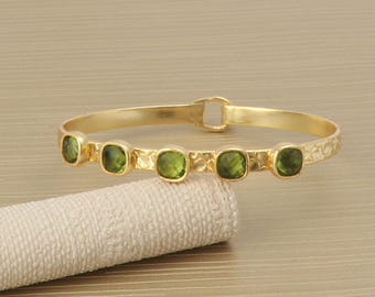 Puede incluir: Un brazalete dorado con cinco piedras preciosas verdes cuadradas. El brazalete tiene un diseño texturizado a lo largo de la banda y un cierre de broche. Las piedras están espaciadas uniformemente.