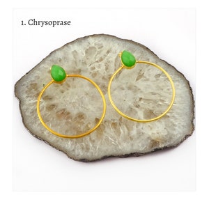 以下が含まれることがあります： ゴールドカラーのフープピアス。上部に涙型の淡い緑色の宝石が付いています。ピアスは、暗い縁取りのある明るい色の楕円形の石の上に展示されています。「1. Chrysoprase」の文字が左上にあります。