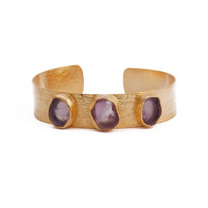 Puede incluir: Brazalete de puño dorado con superficie texturizada. El brazalete presenta tres piedras preciosas moradas, de forma irregular, engarzadas en bisel. El diseño abierto facilita su uso. Una joya elegante.