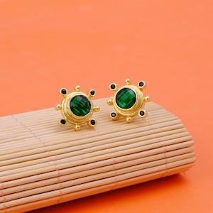 May include: A pair of gold-toned stud earrings. Each earring features a large, round, emerald-green gemstone at the center, surrounded by smaller black accents. The earrings are displayed on a bamboo mat against an orange background.