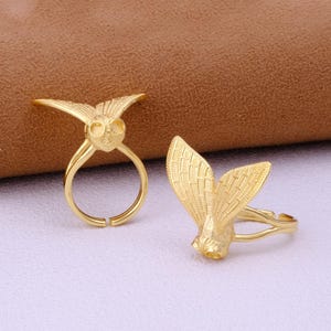 Può includere: Due anelli color oro, uno con un design a gufo e l'altro a farfalla. L'anello a gufo ha un viso dettagliato con grandi occhi e ali. L'anello a farfalla ha ali dettagliate. Entrambi gli anelli sono su una superficie viola chiaro.