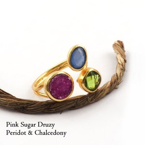 Op de afbeelding: Een goudkleurige ring met drie edelstenen: een roze druzy, een blauwe chalcedoon en een groene peridot. De ring ligt op een witte achtergrond met een bruin touw. De tekst op de afbeelding luidt "Pink Sugar Druzy, Peridot & Chalcedony."