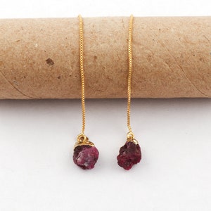 Puede incluir: Un par de pendientes de cadena dorada con gotas de rubí rojo. Los pendientes están hechos con una delicada cadena de caja y presentan piedras de rubí sin pulir.