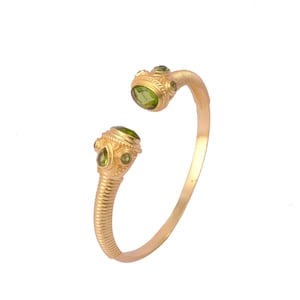 May include: Gold-toned cuff bracelet featuring green gemstone accents. The open-ended design has decorative, textured terminals with oval and round green stones. The bracelet's curved band is smooth, and the terminals have a detailed, ornate design.
